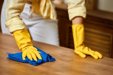 Sarı eldivenli esmer kadın evde mobilya temizlerken bez kullanıyor.