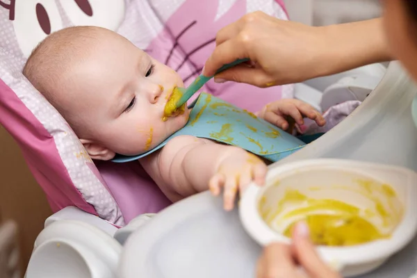 ママは赤ん坊の女の子にベビーチェアのスプーンを与えます 母親は7ヶ月の赤ちゃんにマッシュ野菜をあげ — ストック写真