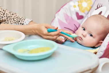 Mom feeds her baby girl with a spoon on baby chair. mother gives food to her baby