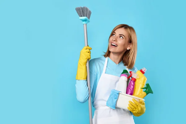 smiling blonde woman in yellow rubber gloves and cleaner apron holding bucket of detergents and broom on blue background.