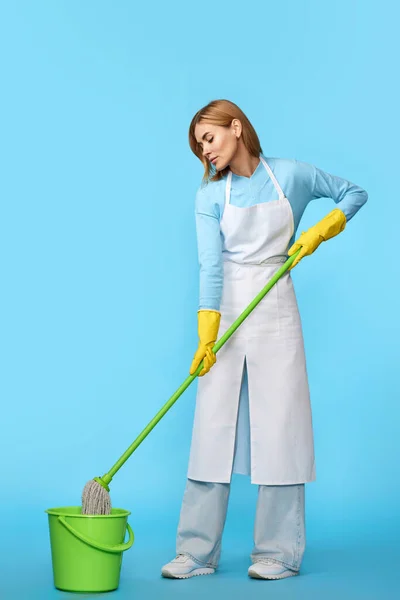 happy cheerful woman in rubber gloves and cleaner apron washing floor with mop on blue background. Full length