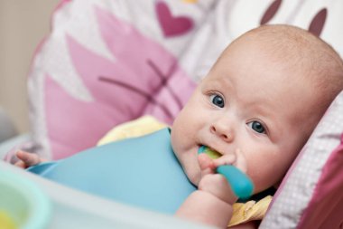 cute baby girl eating with spoon on baby chair in kitchen