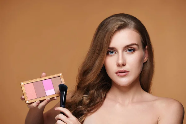 young woman applying blush on cheekbone with make-up brush and holding makeup palette on beige background. contouring