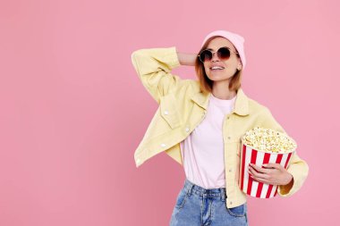 Güneş gözlüklü, şapkalı, film izleyen, elinde pastel pembe arka planda patlamış mısır tutan neşeli genç bir kadın. Boşluğu kopyala