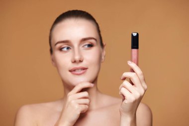 Gülümseyen bir kadın, bej renkli pastel arka planda izole edilmiş çıplak dudak parlatıcısına bakar. dudak parlatıcısına odaklan