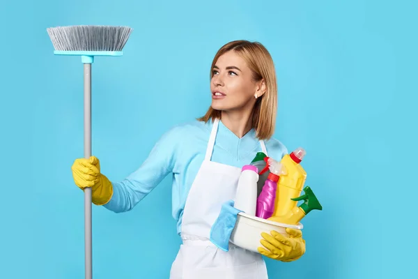 pretty woman in yellow gloves and cleaner apron holding bucket of detergents and looking at broom on blue background. cleaning time