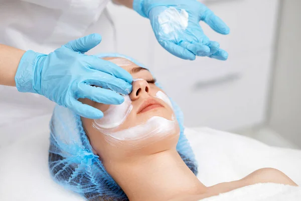 beautician applies the mask to the face of beautiful young woman in the spa salon. facial skin care concept.