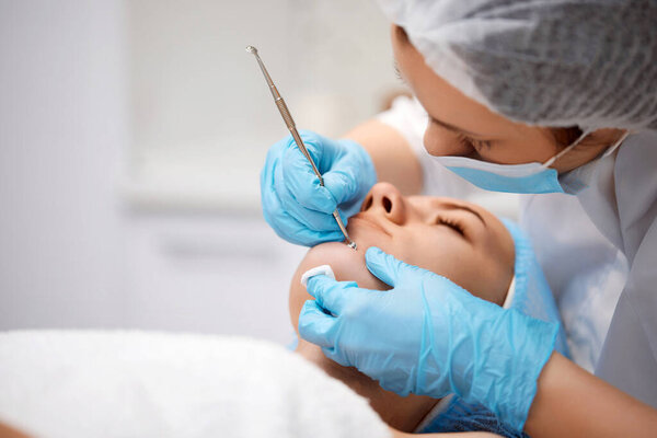 молодая женщина во время механической процедуры очищения лица в клинике красоты. Процедура косметолога для очистки кожи стальным инструментом от угрей и угрей