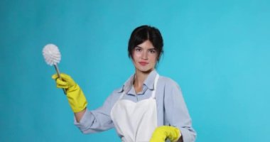 Sarı lastik eldivenli ve temiz önlüklü bir kadın tuvalet fırçası ve mavi arka planda bir şişe deterjan ile dans ediyor..