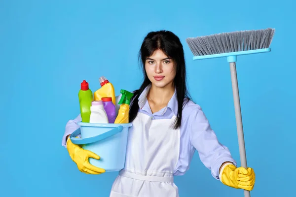 Attractive cheerful woman in yellow rubber gloves and cleaner apron holding bucket with cleaning supplies and broom on blue background.