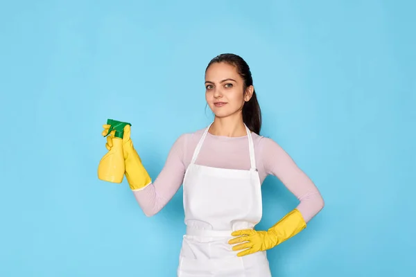 young confident woman in rubber gloves and cleaner apron holding detergent sprayer on blue background.