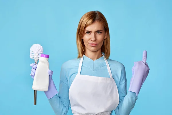 protest woman in rubber gloves and cleaner apron holding toilet brush and bottle of detergent and showing fuck you on blue background.