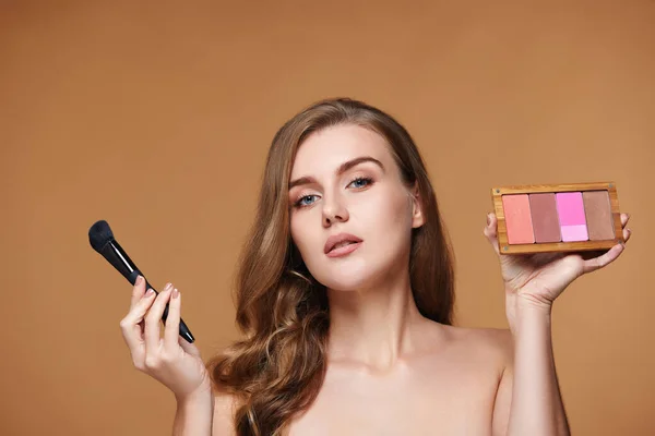 young woman applying blush on cheekbone with make-up brush and holding makeup palette on beige background. contouring