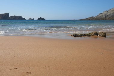 Arka planda kayalıklar olan sakin, kumlu bir sahil.