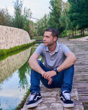 Parkta bir kanalın yanında oturan kot pantolonlu genç bir manken..