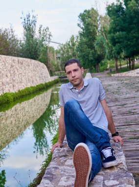 Parkta bir kanalın yanında oturan kot pantolonlu genç bir manken..