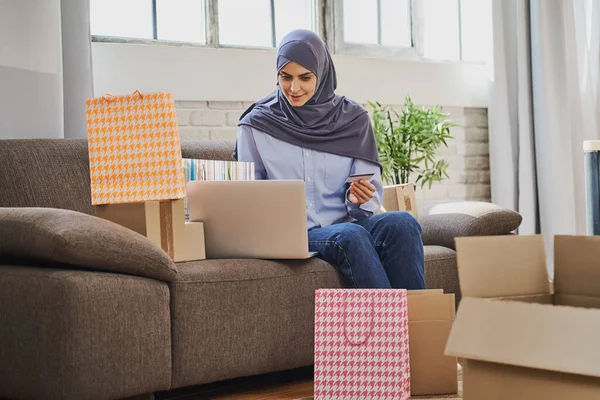 Kanepede oturan neşeli Arap kadın ve hediye almak için dizüstü bilgisayar kullanıyor. Telifsiz Stok Fotoğraflar