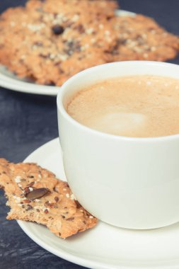 Sütlü bir fincan kahve ve ballı ev yapımı yulaf ezmesi kurabiyeleri ve farklı sağlıklı tohumlar. Lezzetli çıtır tatlı.