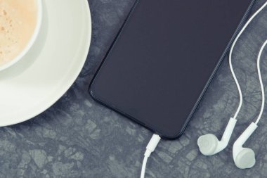 Bağlı kulaklıklı siyah akıllı telefon ve sütlü kahve. Müzikle gevşeme zamanı
