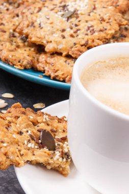 Sütlü bir fincan kahve ve ballı ev yapımı yulaf ezmesi kurabiyeleri ve farklı sağlıklı tohumlar. Lezzetli çıtır tatlı.