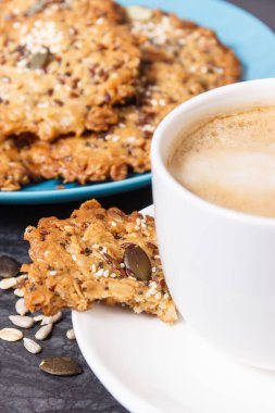 Sütlü bir fincan kahve ve ballı ev yapımı yulaf ezmesi kurabiyeleri ve farklı sağlıklı tohumlar. Lezzetli çıtır tatlı.