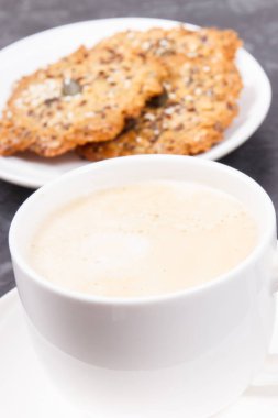 Sütlü bir fincan kahve ve ballı ev yapımı yulaf ezmesi kurabiyeleri ve farklı sağlıklı tohumlar. Lezzetli çıtır tatlı.