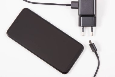 Fişi takılı siyah cep telefonu ve şarj aleti. Akıllı telefon şarj oluyor. Beyaz arkaplan