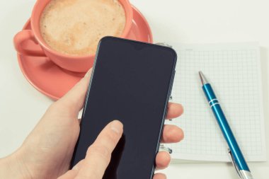 Akıllı telefonlu bir kadın eli, not tutmak için sütlü bir fincan kahve ve not defteri. İş ya da cep telefonuyla rahatlama. Beyaz arkaplan