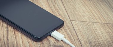Ahşap arka planda şarj kablosu olan siyah akıllı telefon. Cep telefonu şarj kavramı