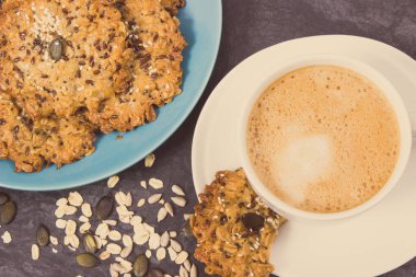 Sütlü bir fincan kahve ve ballı ev yapımı yulaf ezmesi kurabiyeleri ve farklı sağlıklı tohumlar. Lezzetli çıtır tatlı.