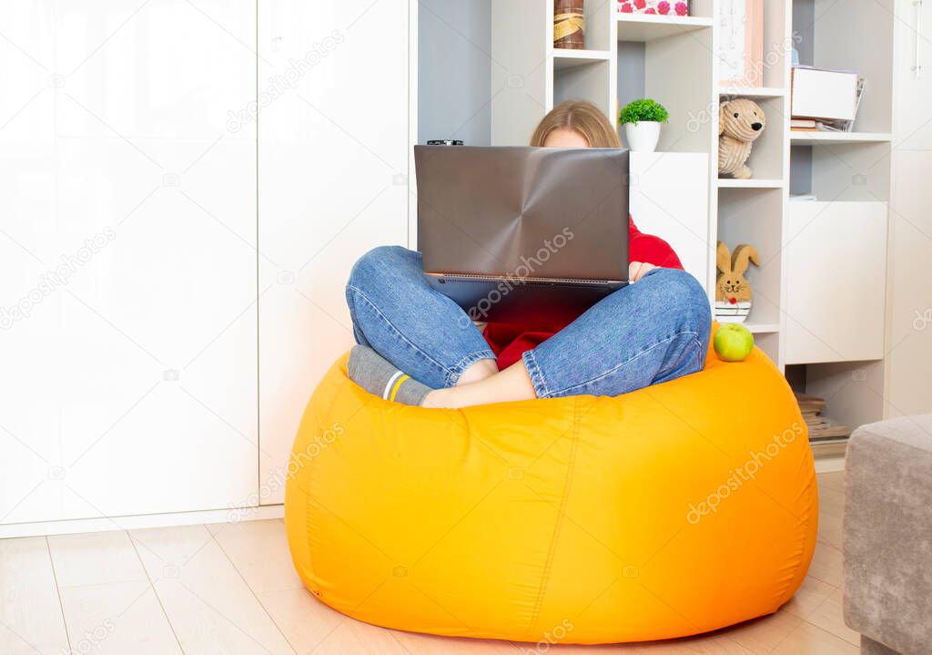 A young woman sits on an orange beanbag chair in the room and works using a laptop at home. Remote work concept, freelance.