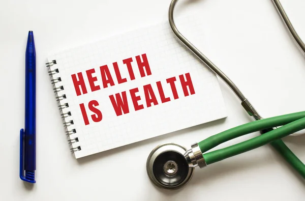 GESUNDHEIT IST WEALTH steht in einem Notizbuch auf einem weißen Tisch neben Stift und Stethoskop. — Stockfoto