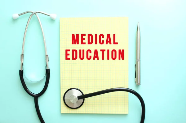 The red text MEDICAL EDUCATION is written in a yellow pad that lies next to the stethoscope on a blue background. Лицензионные Стоковые Фото