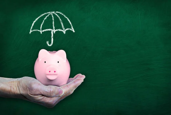 手拿着粉红的小猪银行在黑板前展示粉笔画的伞 雨天储蓄 金融保险 投资或有复制空间的银行业务的概念 — 图库照片