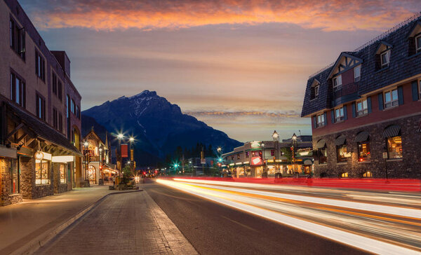 Sunset on Banff Avenue in Banff National Park with Cascade Mountain in the background as light trails from cars lit up the street during night fall.