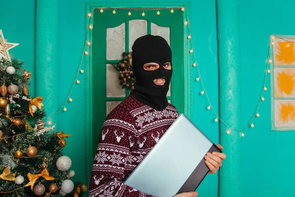 A masked bandit stole a laptop in the new year and escapes. A man in a balaclava holds a computer. theft for christmas