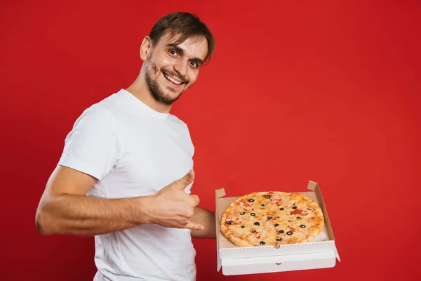 A young man in a white T-shirt holds a box in his hands and eats pizza. Handsome guy on a red background eats fast food.Mock-up.