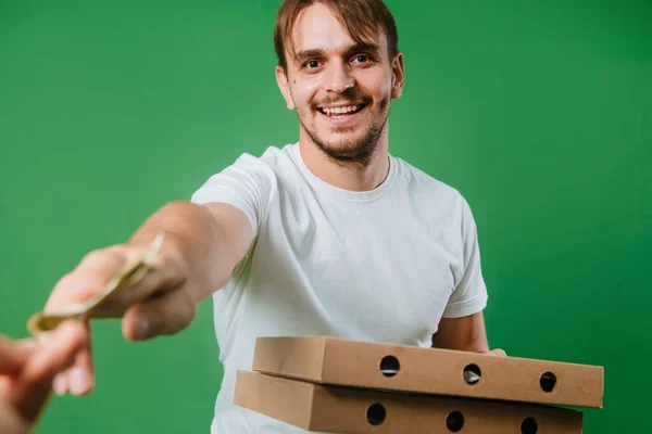 A hand gives money to a young man in a white T-shirt. The guy is holding boxes of pizza on a green background. Photo pizza delivery man in studio