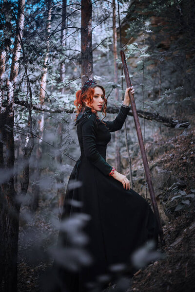Evil witch with a staff in the forest. Slavic witch creates red magic. A young woman in a black dress and a crown embroidered with beads. Halloween costume fantasy