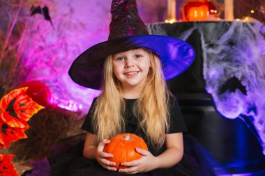 Kasvetli bir arka planda cadı gibi giyinmiş tatlı sarışın kız, elinde bir balkabağı tutuyor. Azizler Günü için dekorasyon. Cadılar Bayramı kostümü. Büyücü şapkalı çocuk.