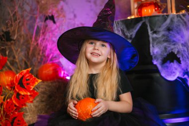 Kasvetli bir arka planda cadı gibi giyinmiş tatlı sarışın kız, elinde bir balkabağı tutuyor. Azizler Günü için dekorasyon. Cadılar Bayramı kostümü. Büyücü şapkalı çocuk.