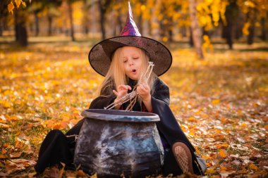 Cadı gibi giyinmiş tatlı sarışın kız sonbaharda parkta iskeletlerle oynuyor. Azizler Günü için dekorasyon. Cadılar Bayramı kostümü. Büyücü şapkalı bir çocuk kazanın yanında oturuyor.