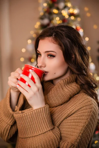 クリスマスツリーとガーランドの背景に暖かいセーターの美しい女の子は 赤いマグカップからコーヒーを飲んでいます クリスマスの装飾を背景にしたモデル — ストック写真