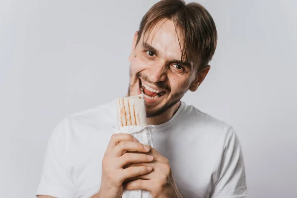 A young man in a clean T-shirt on a white background holds lunch in his hands, shawarma with chicken. The guy is eating fast food.