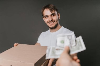 Beyaz tişörtlü, kare pizza kutularıyla gülümseyen genç bir adam gri arka planda duruyor ve para alıyor.