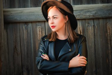 Ahşap bir çitin yanında, kırsal tarzda, şapka ve deri ceketli şık bir siyah kız. Sonbahar resmi.