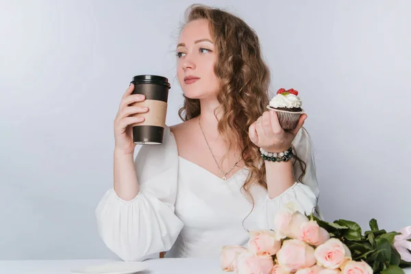 白い背景にカールを持つ美しい若い女性は 彼女の手の中にクリームとベリーとコーヒーや紅茶のプラスチック製のカップケーキを保持しています ケーキで酒を飲む — ストック写真