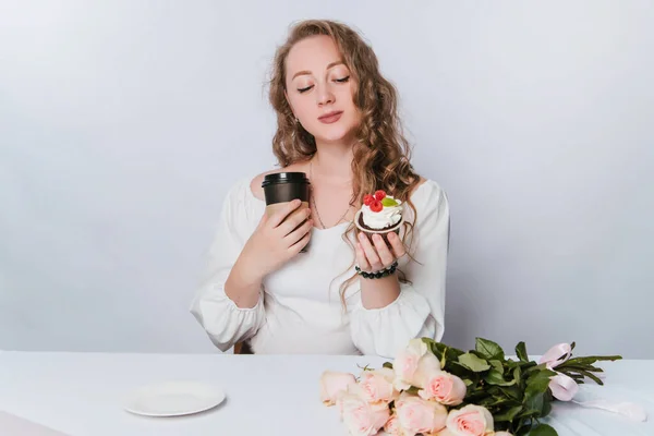 白い背景にカールを持つ美しい若い女性は 彼女の手の中にクリームとベリーとコーヒーや紅茶のプラスチック製のカップケーキを保持しています ケーキで酒を飲む — ストック写真