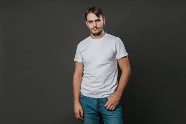Beyaz tişörtlü ve kot pantolonlu yakışıklı bir adam gri arka planda duruyor. Stüdyo fotoğrafı. Model.
