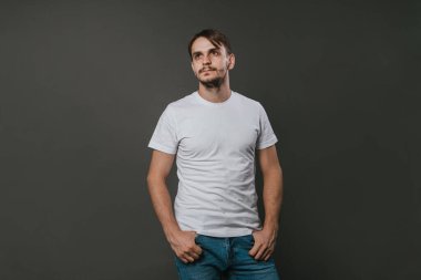 Beyaz tişörtlü ve kot pantolonlu yakışıklı bir adam gri arka planda duruyor. Stüdyo fotoğrafı. Model.